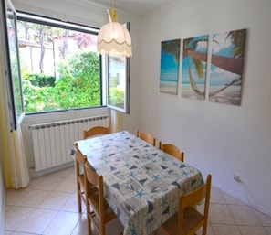 A dining area with a table and chairs, a window with a view of greenery, a radiator, and a wall with two paintings. - Holiday Villa With Garden (Lignano Sabbiadoro)