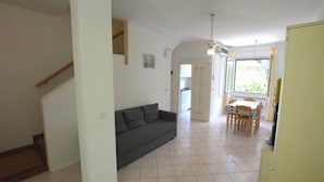 A living room with a dark grey sofa, a dining area with a table and chairs, and a staircase leading to another level. - Holiday Villa With Garden (Lignano Sabbiadoro)
