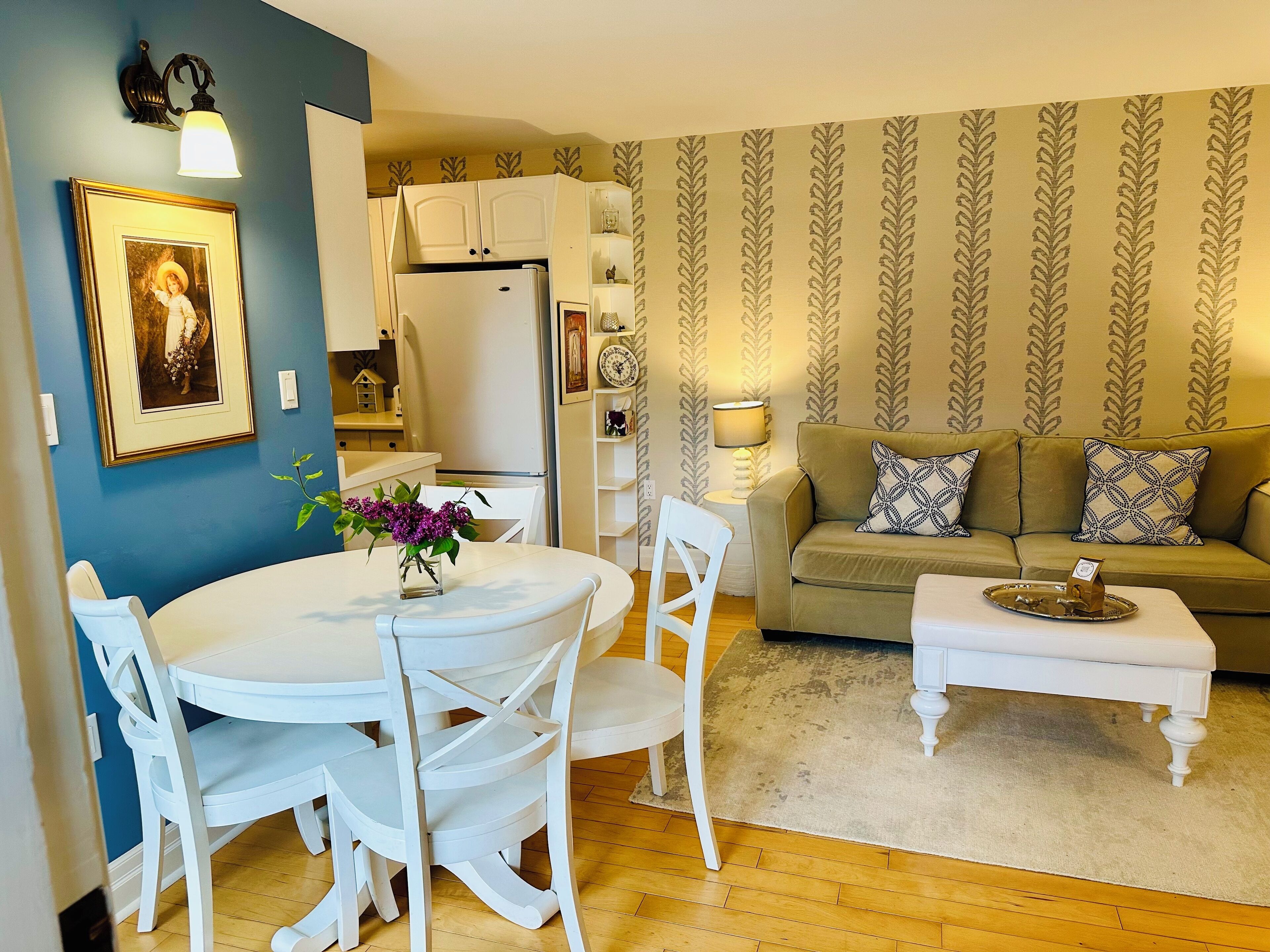 Un salón con una mesa redonda, sillas blancas, un sofá y una mesa de centro. Hay una zona de cocina con armarios blancos y un refrigerador.
