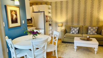 Un salón con una mesa redonda, sillas blancas, un sofá y una mesa de centro. Hay una zona de cocina con armarios blancos y un refrigerador.