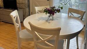 Un comedor con una mesa redonda, sillas blancas y un florero con flores. Se observa una pared de piedra y una puerta con paneles de vidrio.