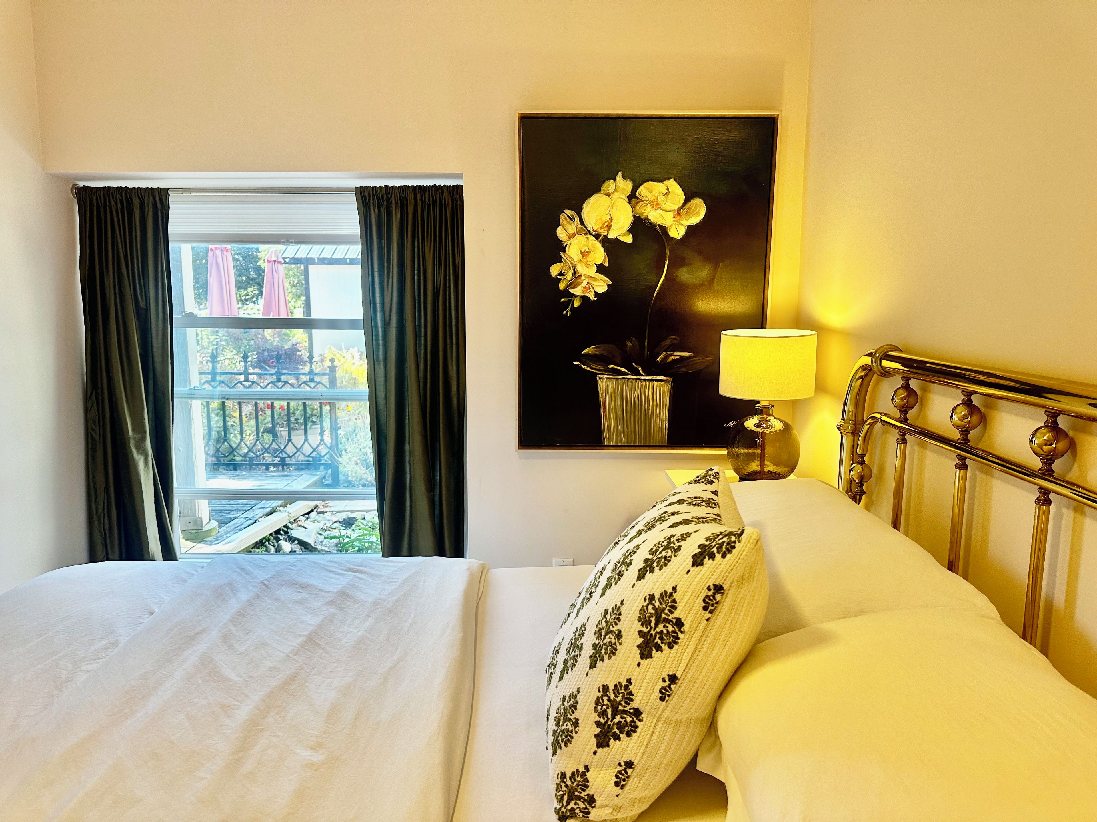 Habitació d'hotel amb un llit, una finestra amb cortines, un quadre de flors i una làmpada de nit.