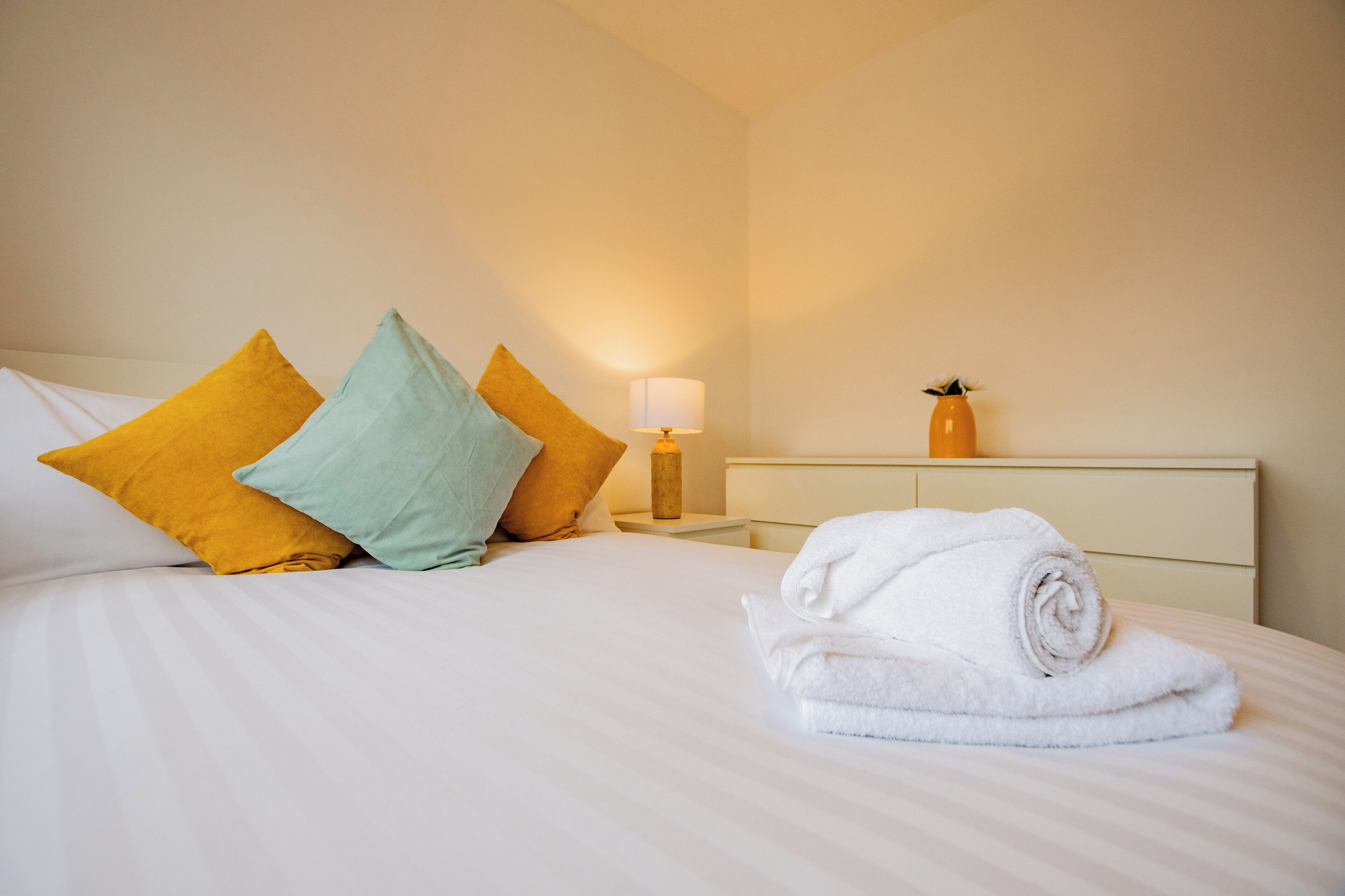 Un lit bien fait, avec des draps blancs, une serviette blanche enroulée et trois oreillers aux couleurs jaune et verte.