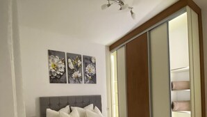 A bedroom with a grey upholstered headboard, a bed with white linens and two pillows, a bedside table with a lamp, and a sliding door closet.