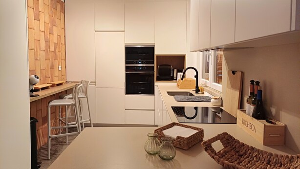 Een moderne keuken met een ingebouwde oven, een magnetron en een spoelbak. Twee krukken en een tafel met een geweven mand en glazen vazen.