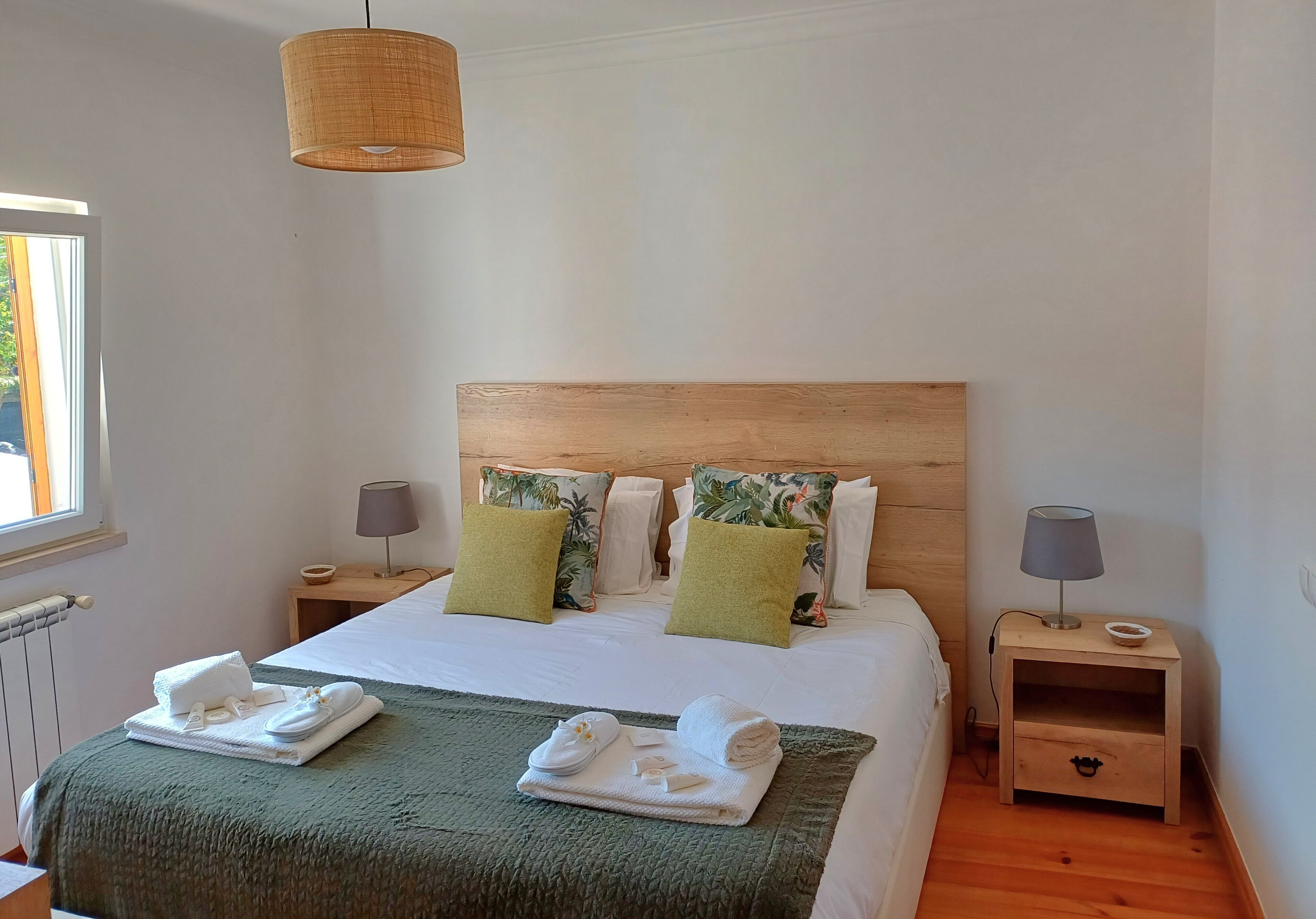 A neatly made bed with a green blanket, two pillows, and a headboard. There are two bedside tables with lamps and a small drawer.