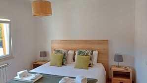 Een net opgemaakt bed met een groene deken, twee kussens en een hoofdeinde. Er staan twee nachtkastjes met lampen en een klein lade.