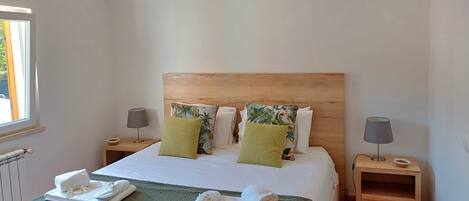 A neatly made bed with a green blanket, two pillows, and a headboard. There are two bedside tables with lamps and a small drawer.