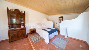 A bedroom with a wooden cabinet, a bed with white bedding, a nightstand, a bedside table with a lamp, a bathroom with a sink and mirror, and a tiled floor. - Kochab Alentejo - Studio House, Sleeps 2 (Gáfete)