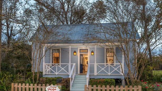 A small, single-story house with a porch, a wooden fence, and a sign indicating it is a country garden cottage.