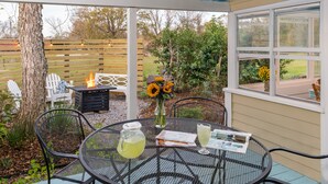 Eine überdachte Terrasse mit einem runden Metalltisch, Stühlen, einer Feuerstelle und Blick auf den Garten.