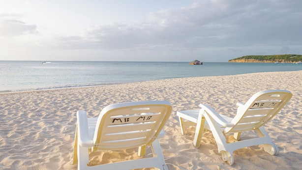 To hvide strandstole med "MB MB" på ryglænet, placeret på en sandstrand med et roligt hav og en båd i det fjerne.