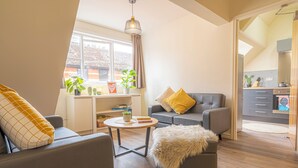 Een moderne woonkamer met een bank, fauteuil en een kleine tafel. Er is een raam met planten en uitzicht op buiten.