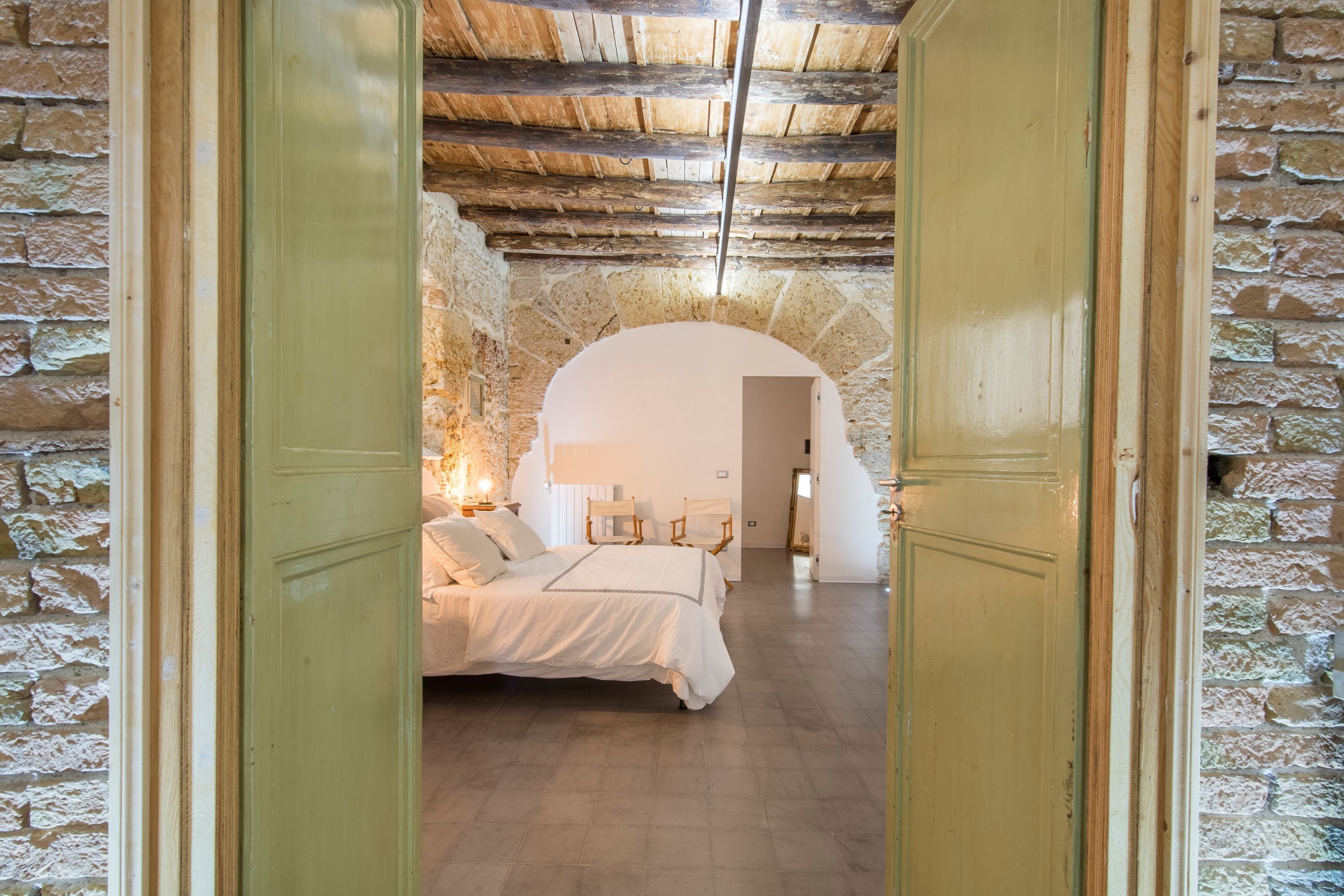 A bedroom with a stone wall, wooden ceiling beams, a bed with white linens, and a nightstand with a lamp.