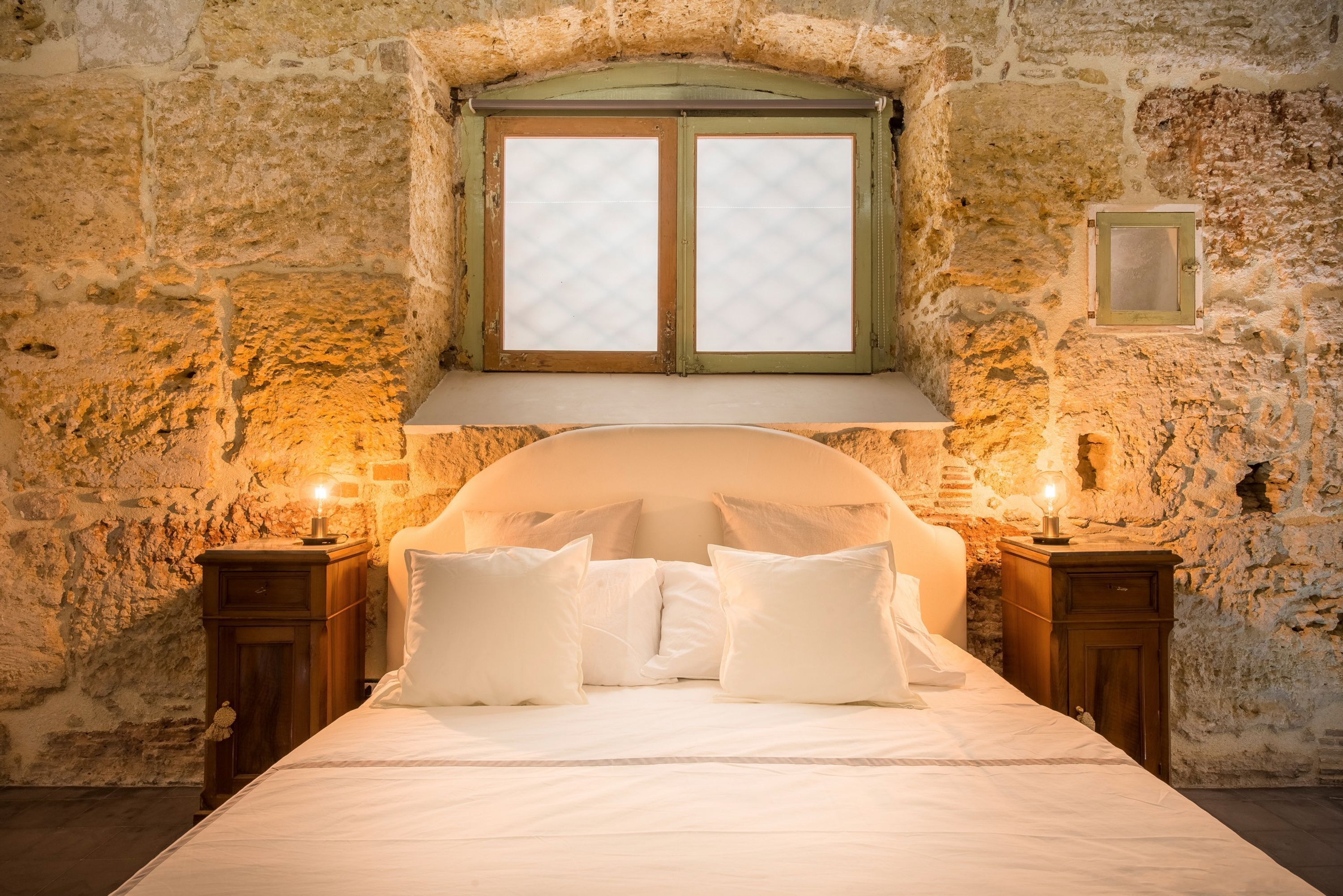 A bedroom with a stone wall, a wooden headboard, a window with a green frame, and two bedside tables with lamps.