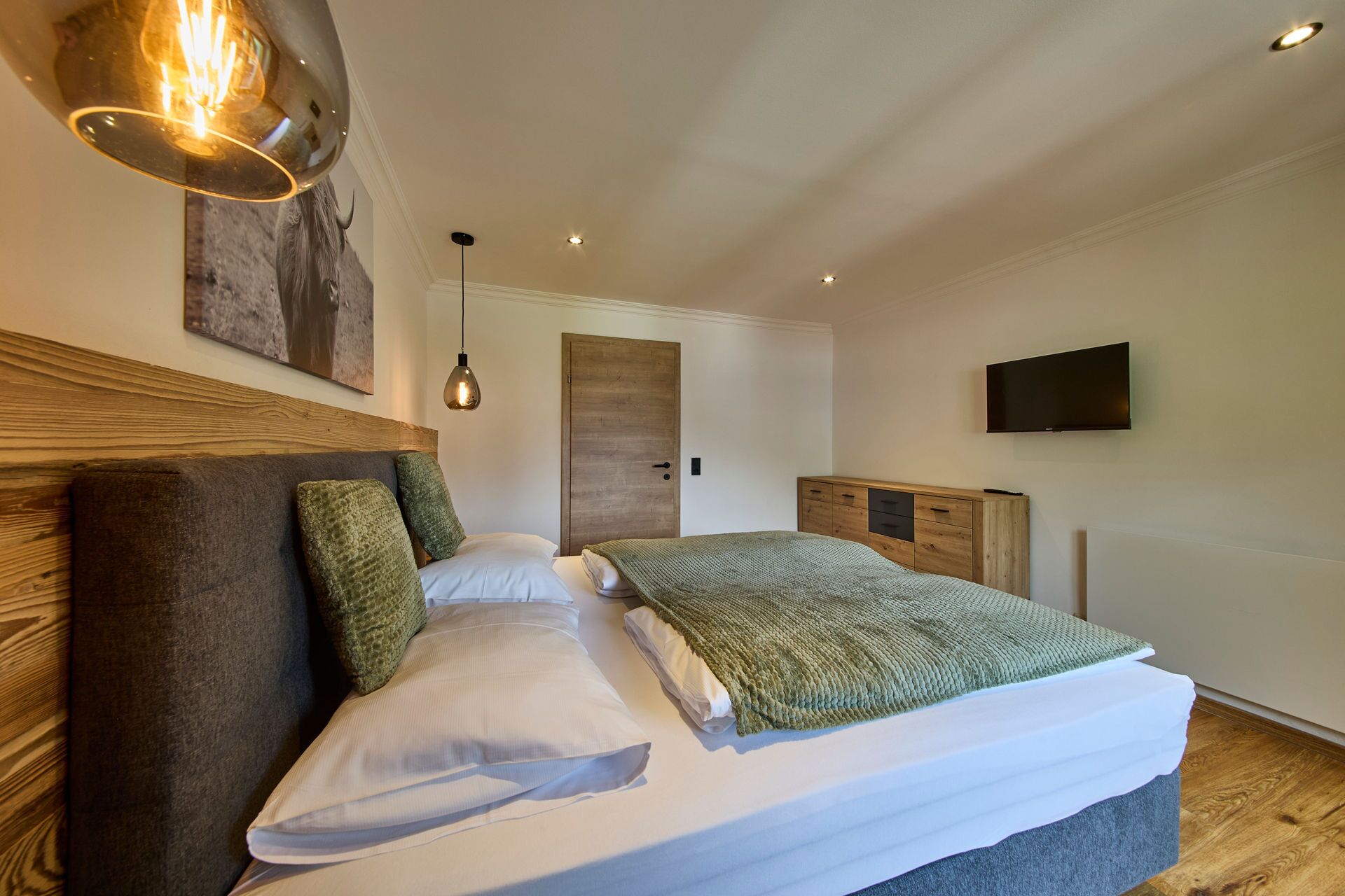 Ein Schlafzimmer mit einem hölzernen Kopfende, einem Bett mit weißer und grüner Bettwäsche, einem an der Wand befestigten Fernseher und einer hölzernen Kommode.