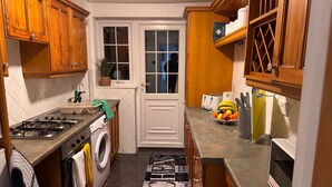 Une cuisine compacte avec des meubles en bois, une cuisinière à gaz, un lave-linge et un panier de fruits posé sur le plan de travail.