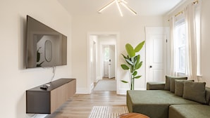 Een moderne woonkamer met een flatscreen televisie, een houten console, een groene bank en een grote plant.