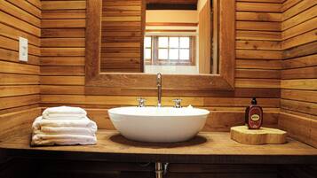 A wooden cabin-style bathroom with a large oval sink, two faucets, and a mirror above it. There are towels stacked on the counter and a bottle of soap.