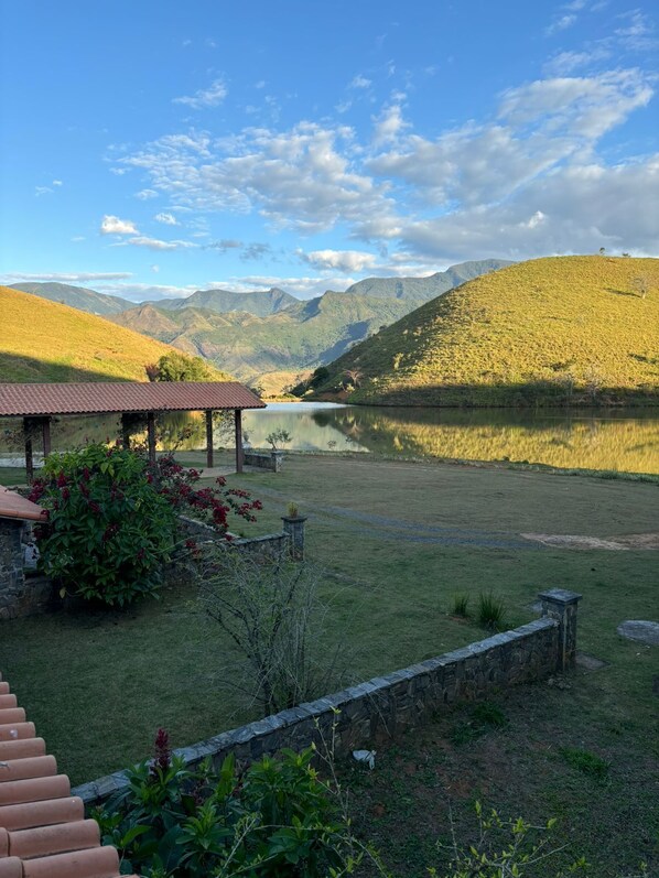 A serene lakeside setting with a stone structure and a red roof, surrounded by lush greenery and distant mountains.