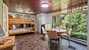 A cozy room with a bunk bed, a dining table with chairs, a kitchen area, and large windows offering a view of greenery.