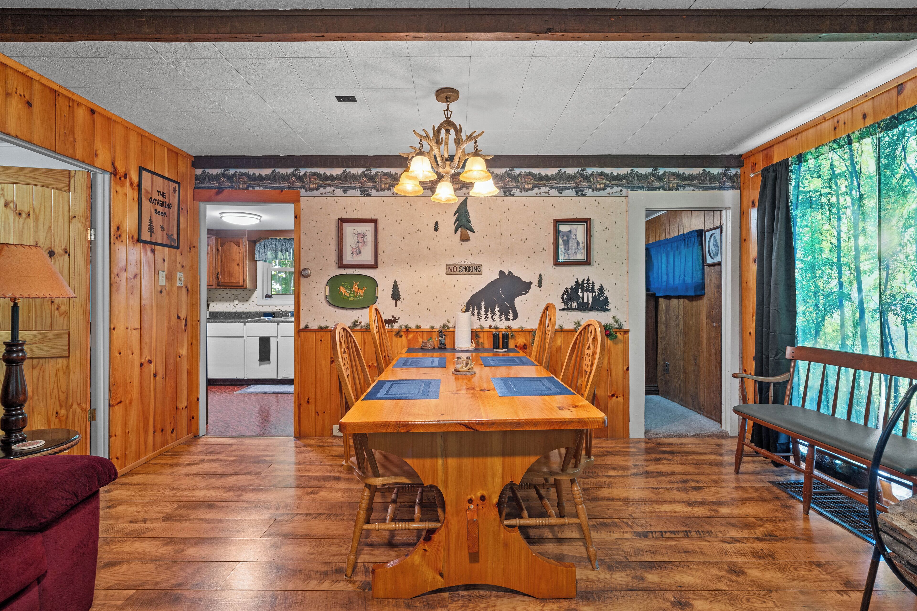 Une salle à manger en bois, avec une grande table et des chaises, un lustre et une vue sur l’extérieur par une fenêtre.