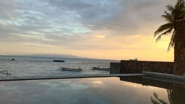 A swimming pool with a view of the sea and boats, a palm tree, and a stone wall. - Utama Villas Beach Front Candidasa (Bali)