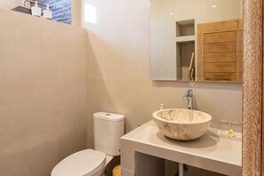 Un baño con inodoro blanco, lavamanos de mármol y un mueble de madera sobre el espejo.
