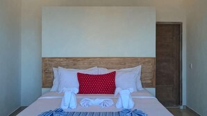 A neatly made bed with white and red pillows, a wooden headboard, and a pair of towels on the bed. - Utama Villas Beach Front Candidasa (Bali)
