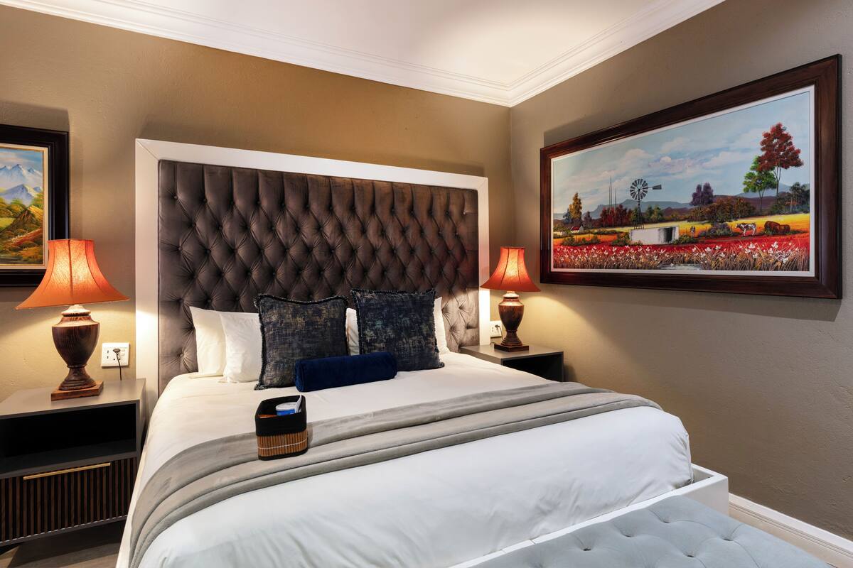 A bedroom with a tufted headboard, a large painting, two bedside lamps, and a bedside table with a lamp.