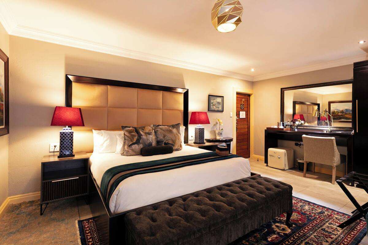 A hotel room with a large bed, two bedside tables with lamps, a desk with a chair, a mirror, and a small side table with a vase.