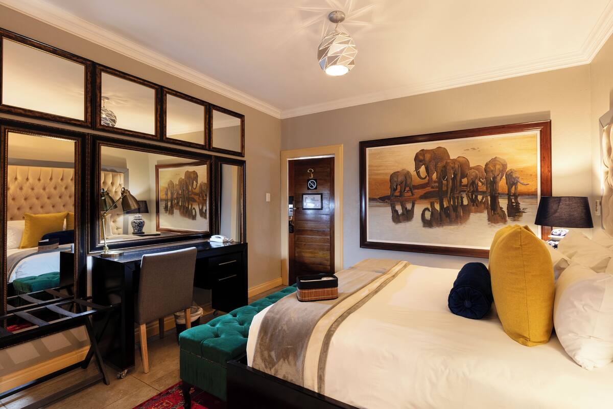 A hotel room with a bed, a desk, a chair, a mirror, and a painting on the wall.