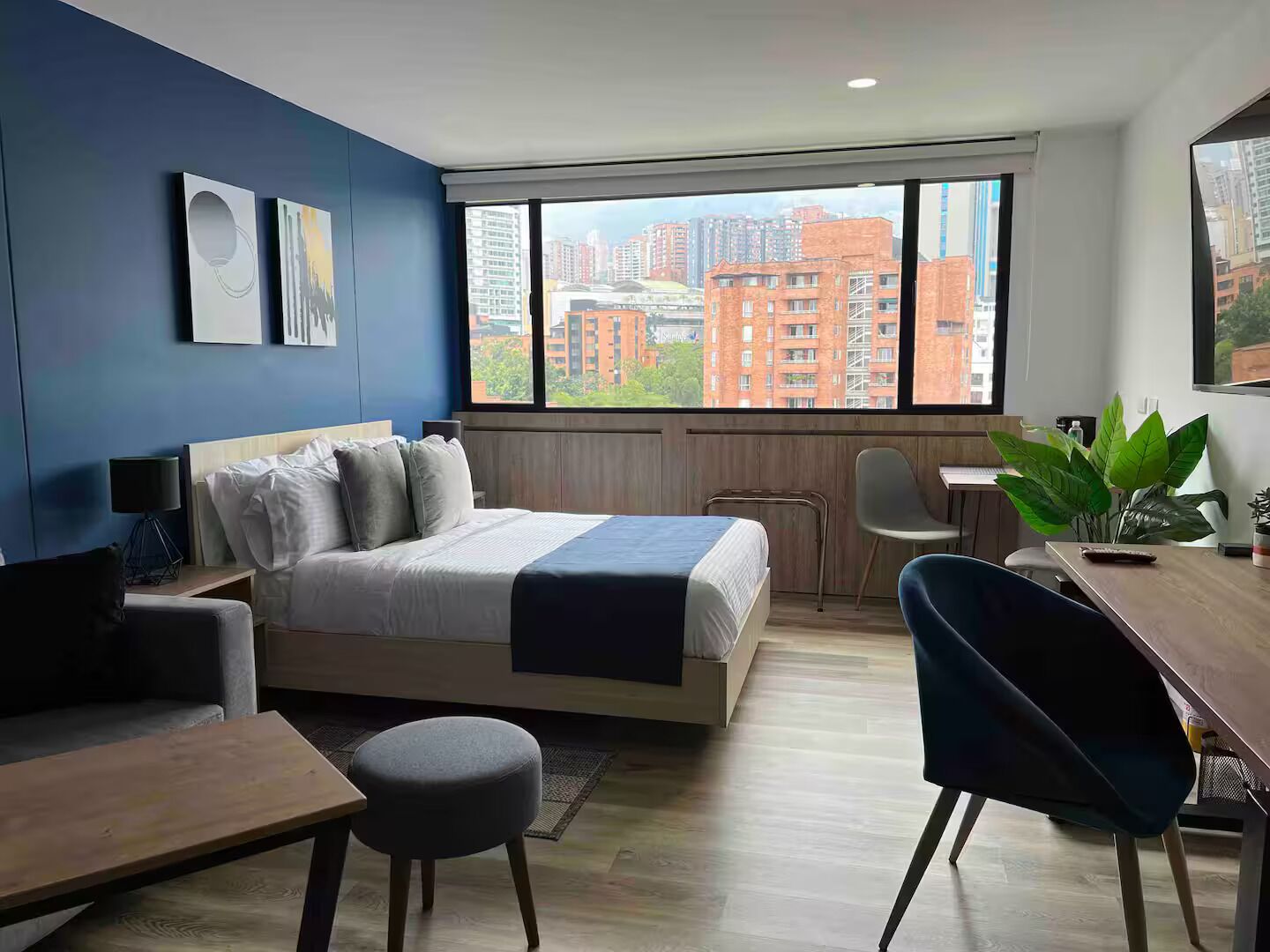 A modern hotel room with a large bed, a desk, a chair, and a view of city buildings.