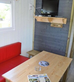 A room with a wooden table, a red sofa, a small wooden stool, a television mounted on a wooden shelf, and a window with a view of greenery outside. - ZEcamping 4 étoiles familial piscine chauffée locatif BBQ clim 36m2 (Secondigny)