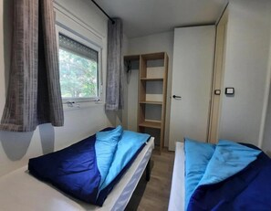 Une petite pièce compacte comprenant deux lits simples, une fenêtre avec des rideaux, une étagère intégrée et une armoire.