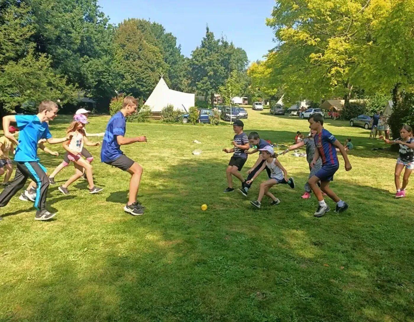 Un grupo de personas jugando a un juego en un campo cubierto de césped, con árboles y tiendas de campaña al fondo.