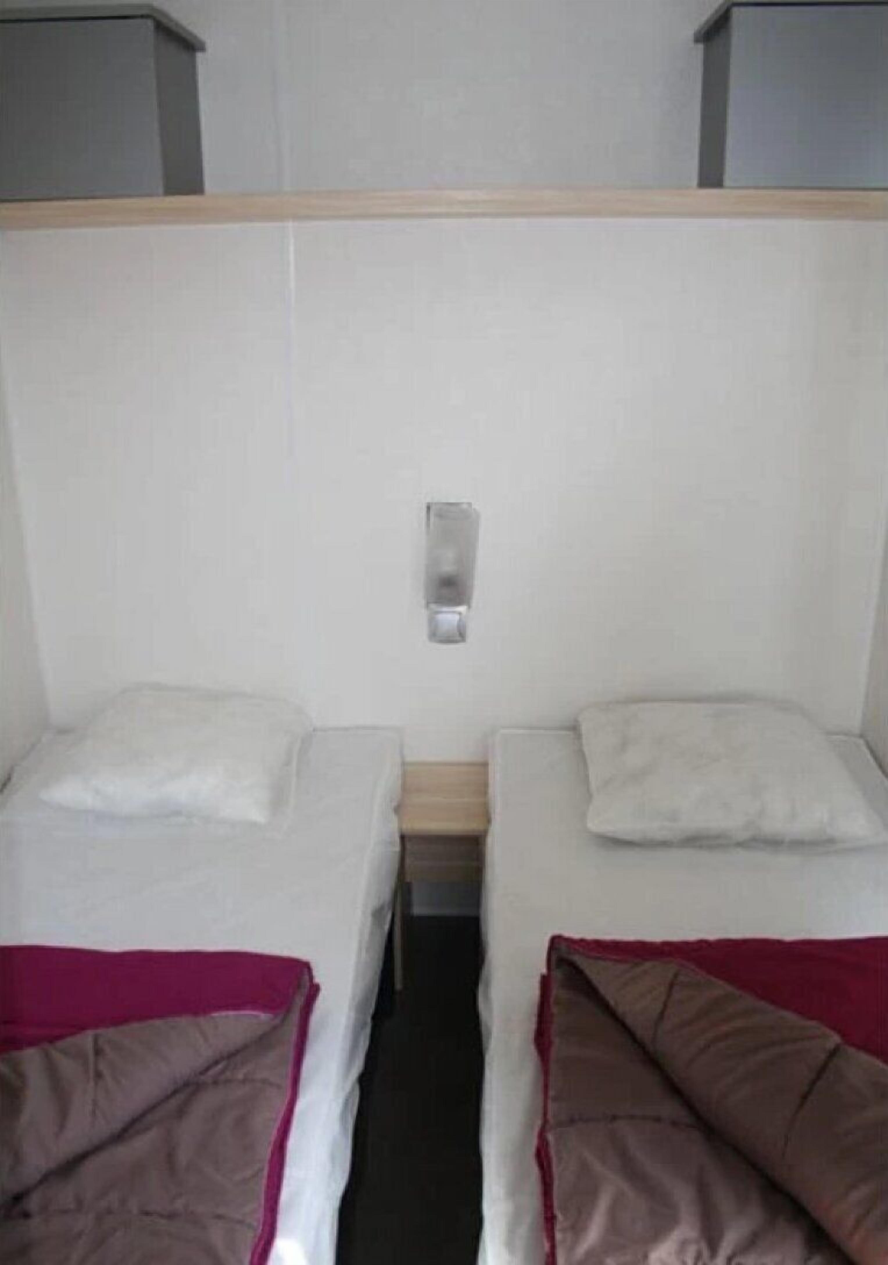 A small, compact room with two single beds, each with a pillow and a blanket. There is a small shelf above the beds and a light switch on the wall.