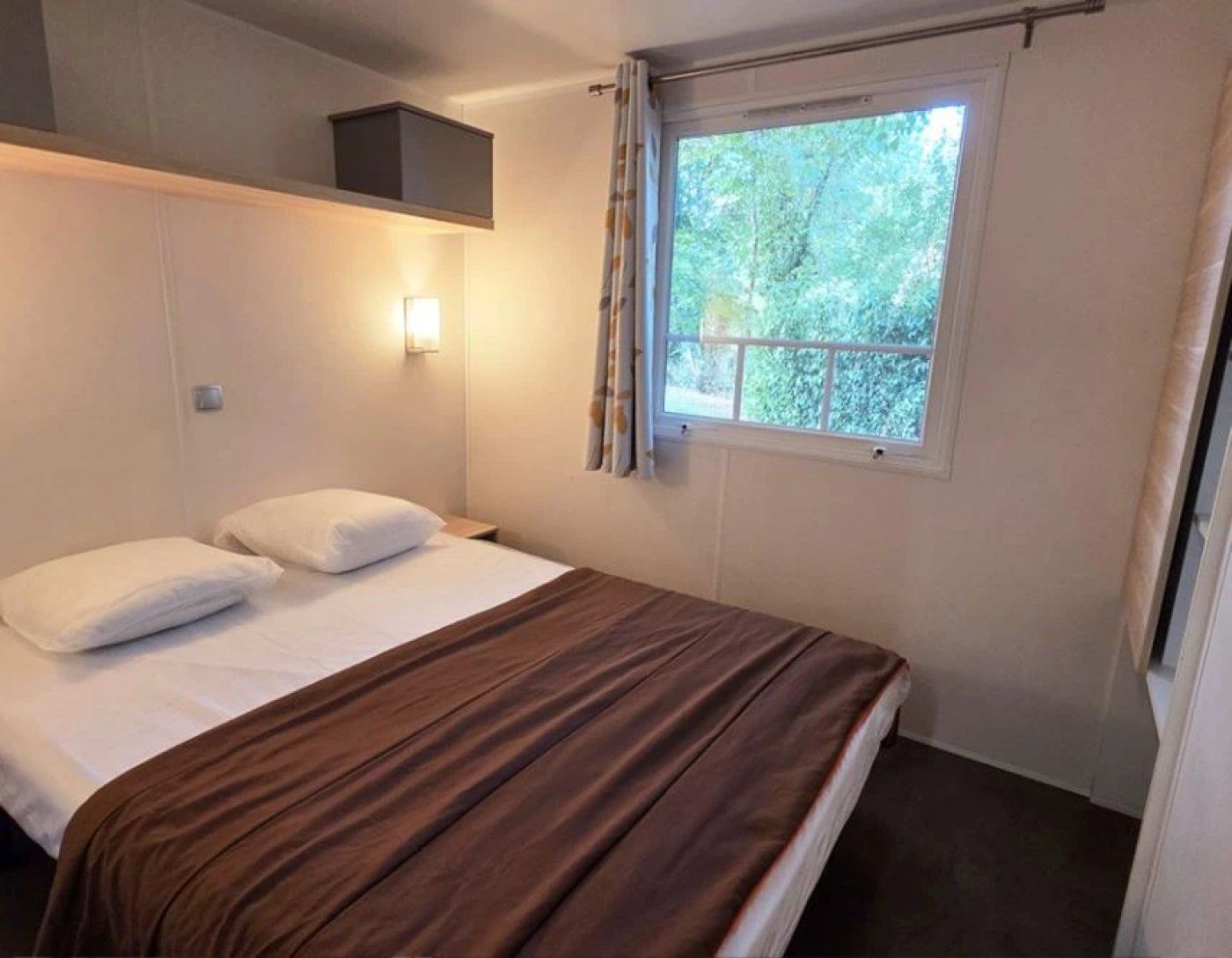 A single bed with a brown headboard, a bedside lamp, and a window with a view of trees.