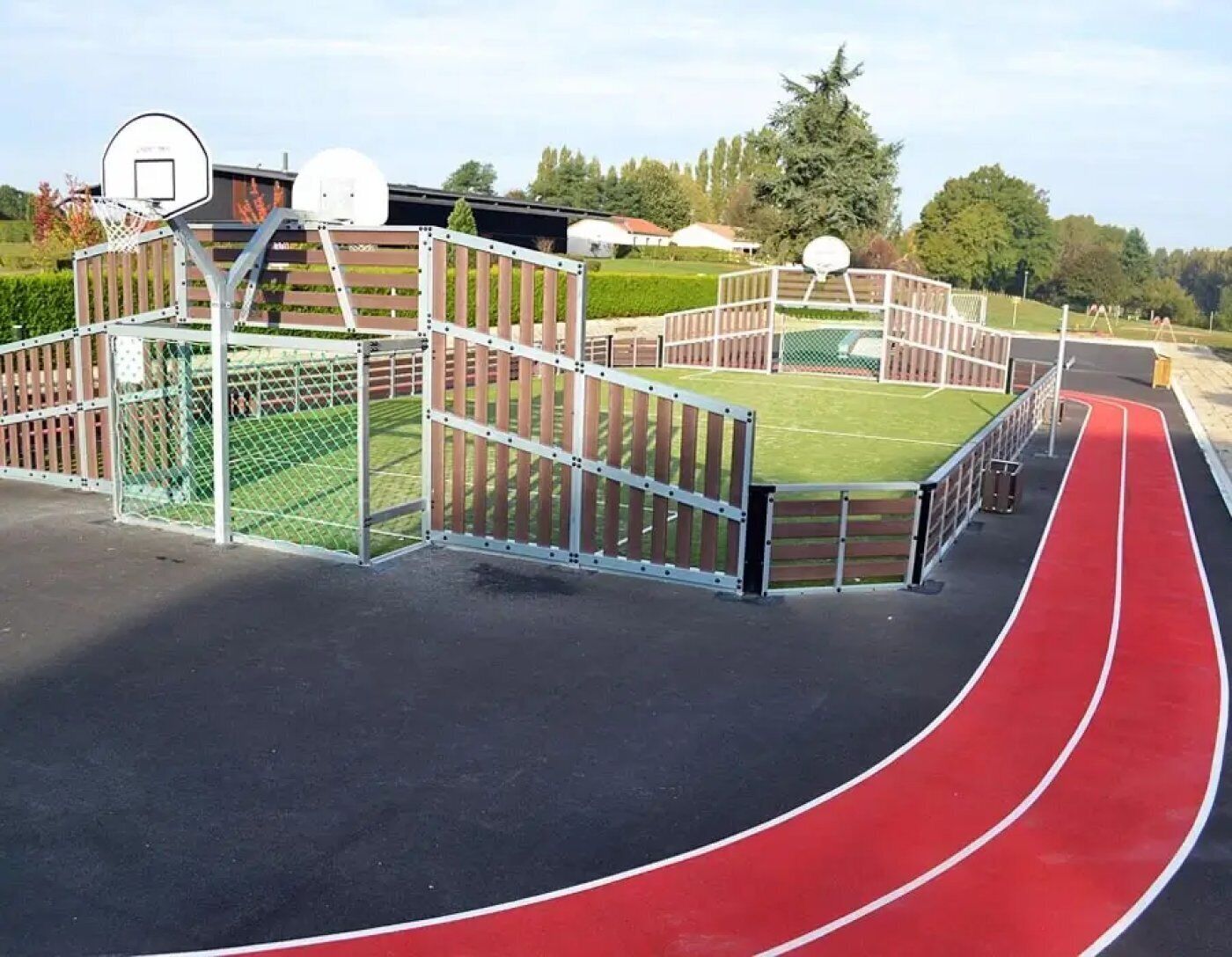 Une piste d’athlétisme à revêtement rouge, un panier de basketball et une zone délimitée par une clôture, avec un terrain vert à l’intérieur.