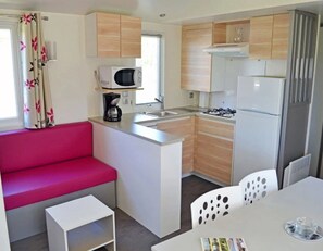 Een kleine keuken met een magnetron, fornuis en koelkast. Er is een zithoek met een rode bank en een witte tafel met stoelen.