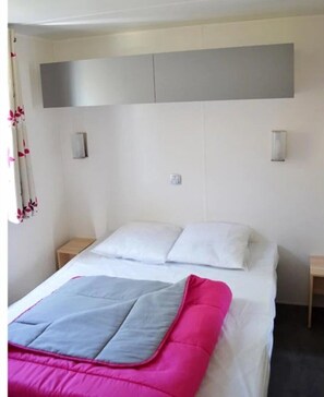 Een netjes opgemaakt bed met een roze en grijze deken, witte kussens en een hoofdbord met een grijze paneel. Er staan twee nachtkastjes met lampen bij, en er is een raam met een gordijn.