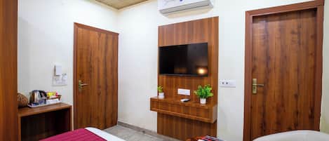 A hotel room with a bed, a TV mounted on a wooden panel, a desk with a kettle, and a chair.
