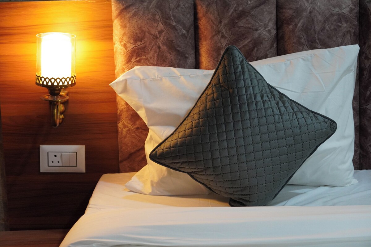A neatly made bed with white linens and a quilted pillow, a wall-mounted light fixture, and a power outlet on a wooden headboard.