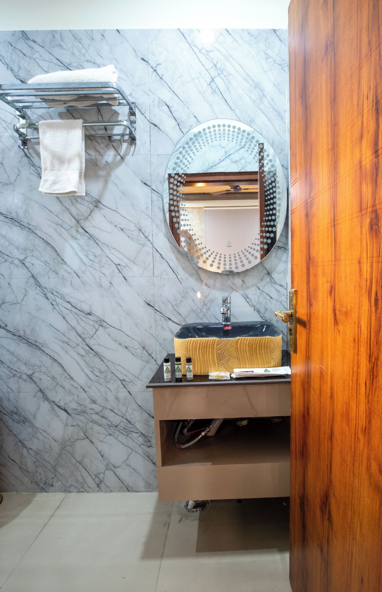 A bathroom with a marble wall, a round mirror, a towel rack, and a wooden door.