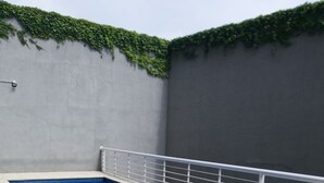 Una piscina con una escalera metálica, rodeada por un suelo de baldosas y una pared gris con follaje verde.