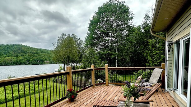 A wooden deck with a table and chairs, overlooking a lake and surrounded by greenery.