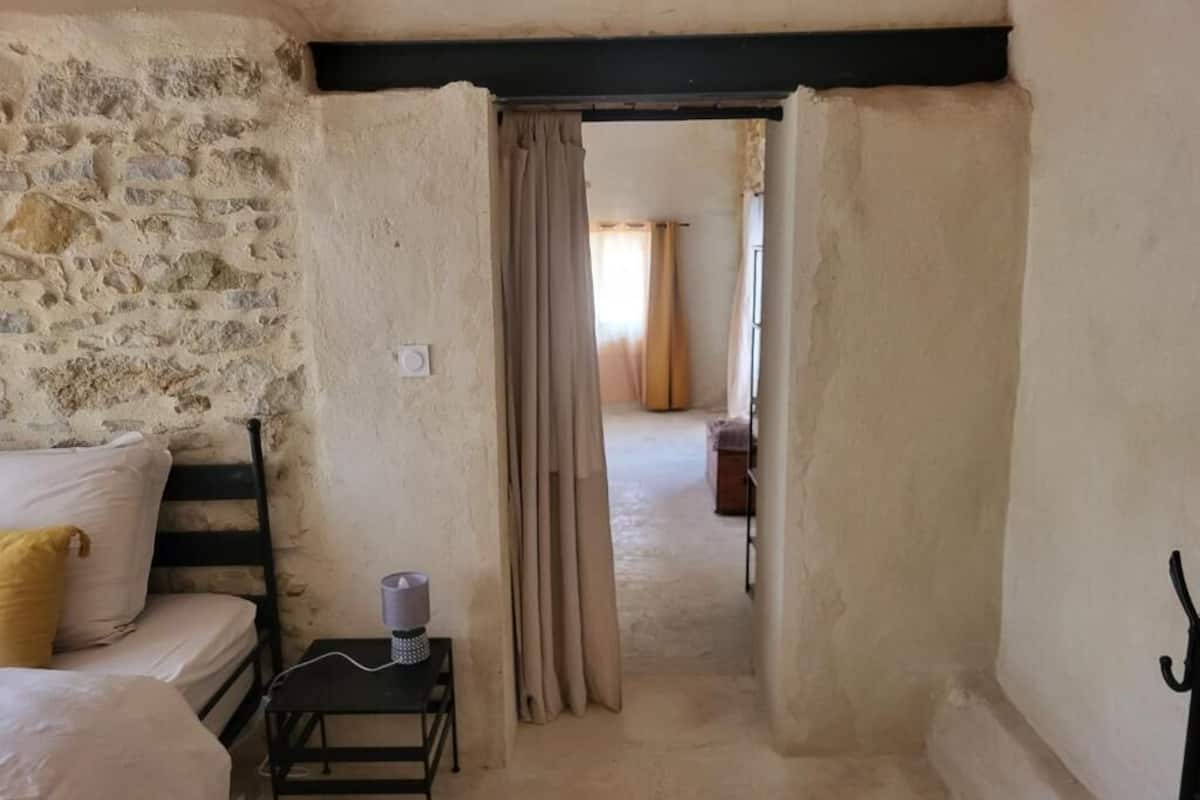 A room with a stone wall, a bed with a white and yellow pillow, a small table with a candle, and a doorway leading to another room.
