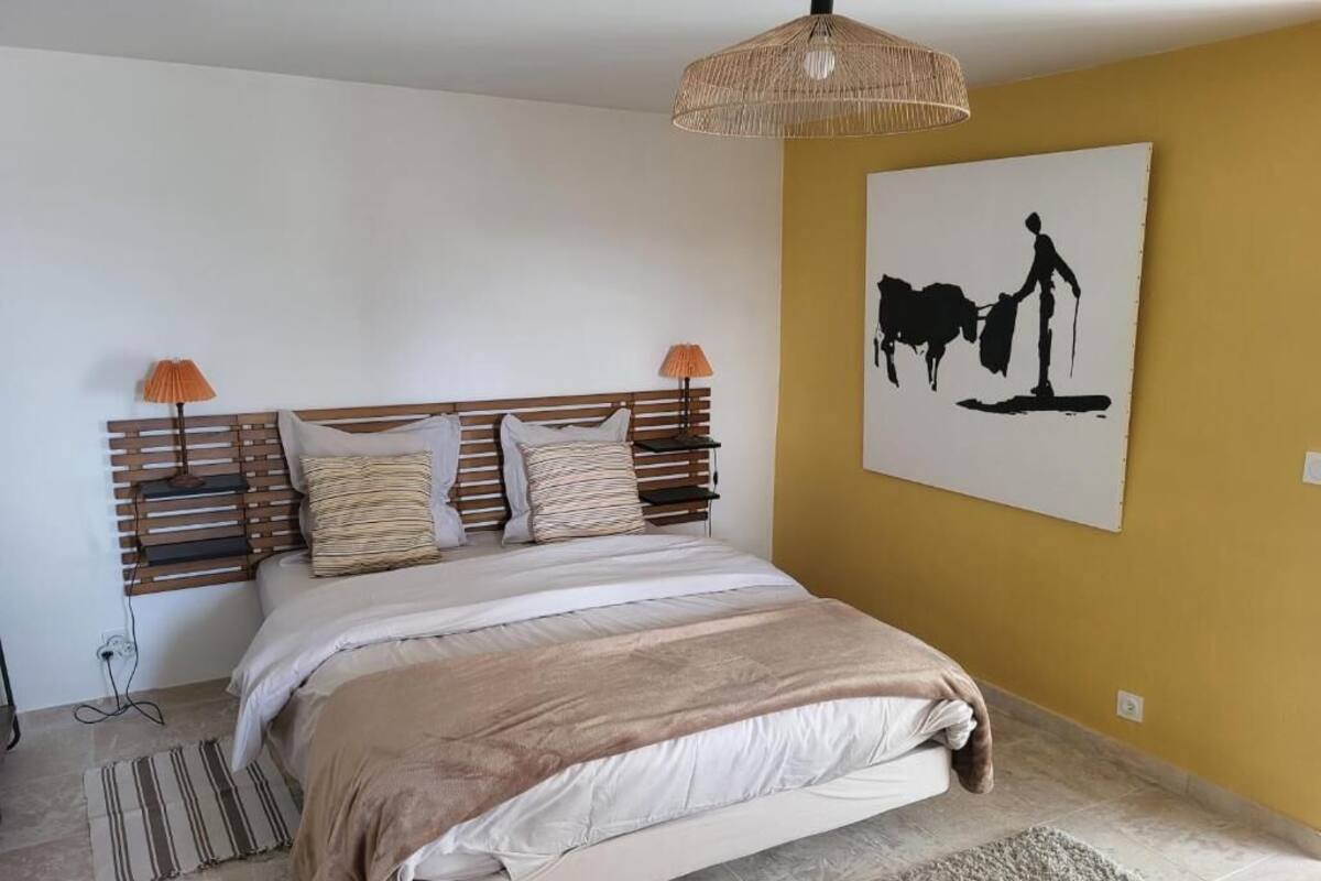 A bedroom with a wooden headboard, a bed with striped bedding, two bedside lamps, a woven light fixture, and a wall-mounted artwork of a cow.
