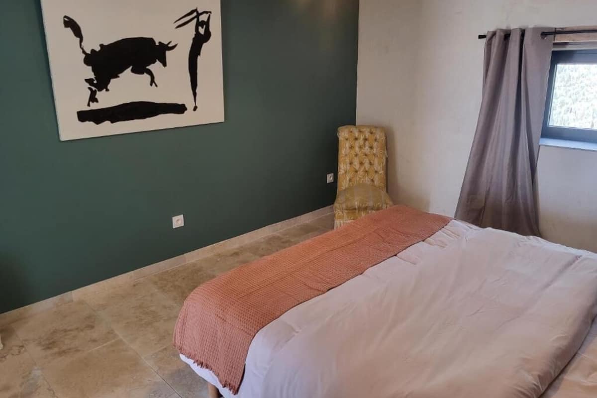 A bedroom with a bed, a chair, a window with curtains, and a wall painting.