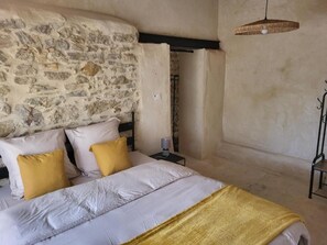 A bedroom with a stone wall, a bed with white and yellow bedding, a bedside table with a lamp, and a hanging light fixture. - bastide de la madeleine (Pougnadoresse)